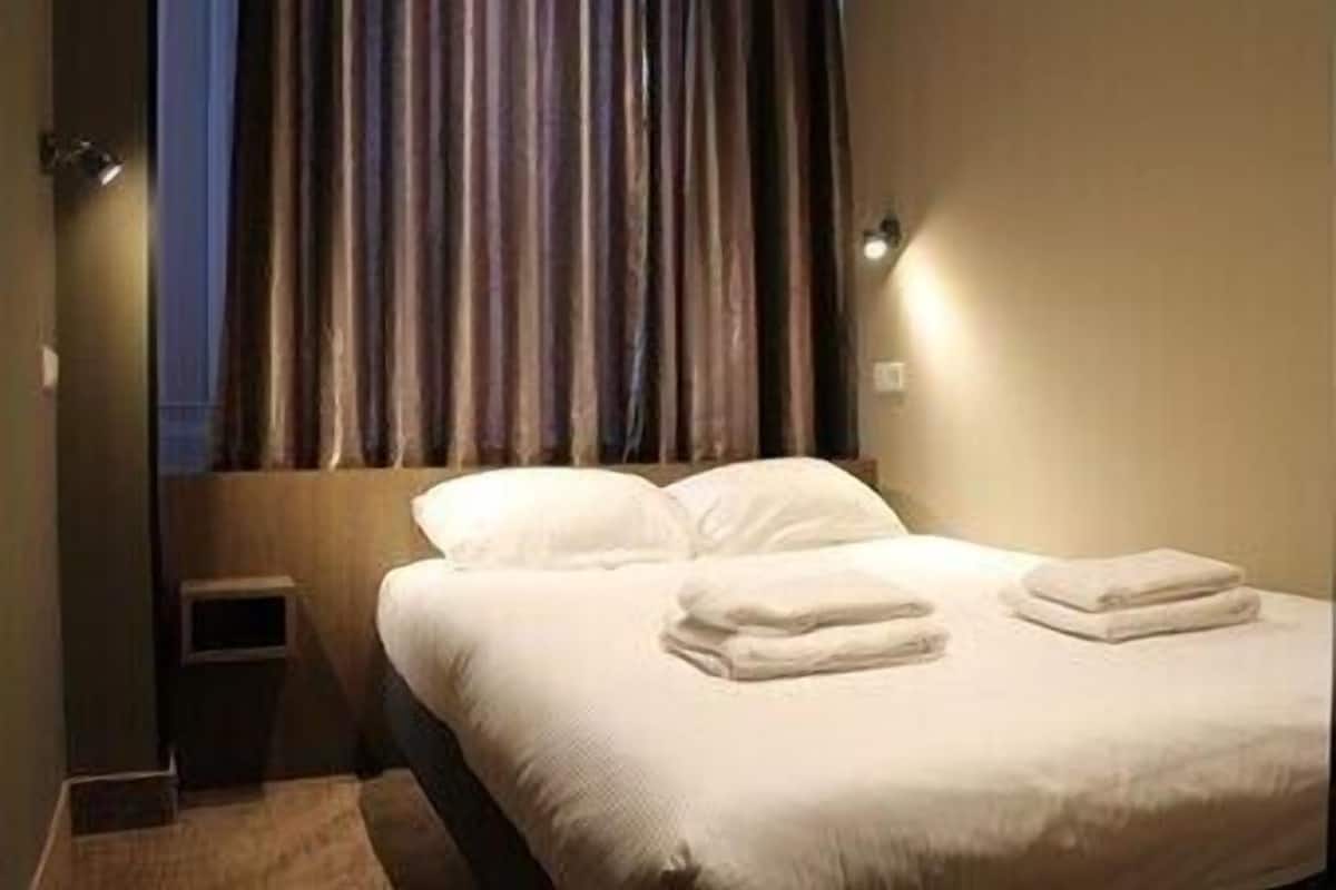 A hotel room with a bed, curtains, a small bedside table, and a wall-mounted light.