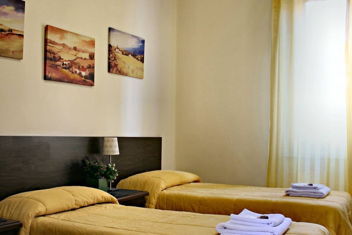 A hotel room with two beds, a nightstand, a lamp, and three landscape paintings on the wall.