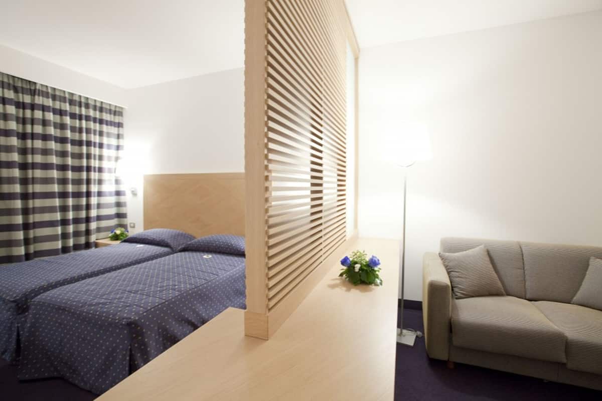 Una habitación de hotel con cama, sofá, un separador de madera y una pequeña mesa con una flor.