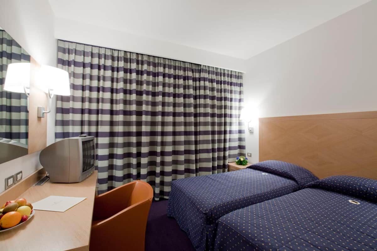 Habitación de hotel con cortina a cuadros, un escritorio con lámpara, un televisor y dos camas con ropa de cama azul.