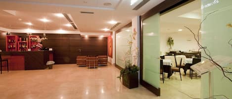 A hotel lobby with a reception desk, seating area, and a dining section visible through a glass partition.
