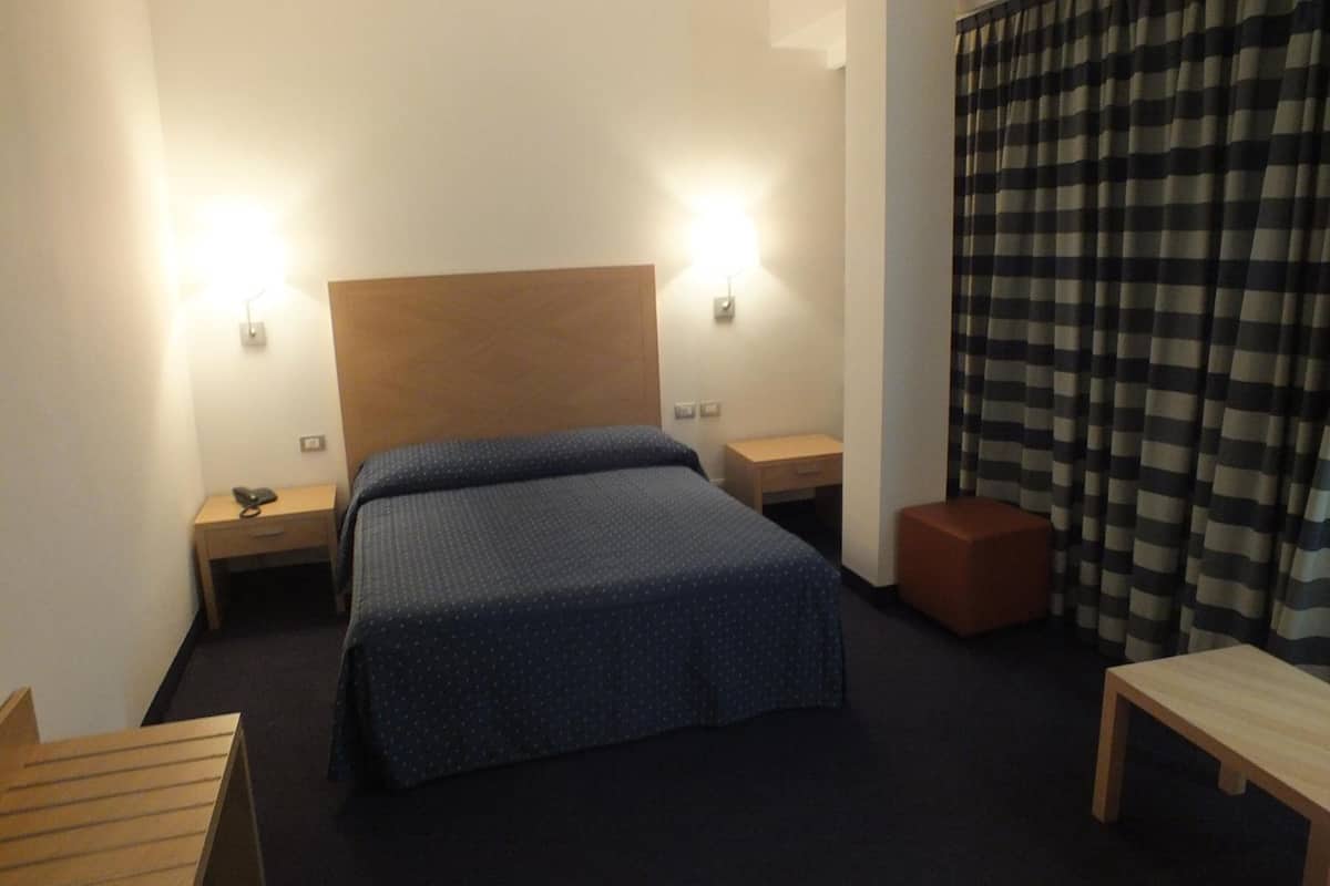 Una habitación de hotel con una cama individual, dos mesitas de noche, un banco de madera y una cortina a cuadros.