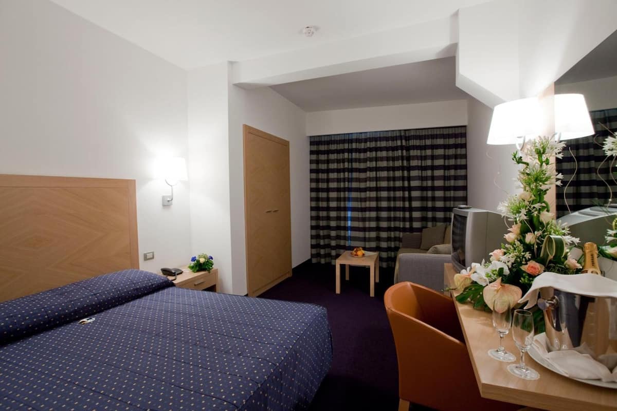 Habitación de hotel con cama, escritorio con lámpara, una silla, una mesita y ventana con cortinas.