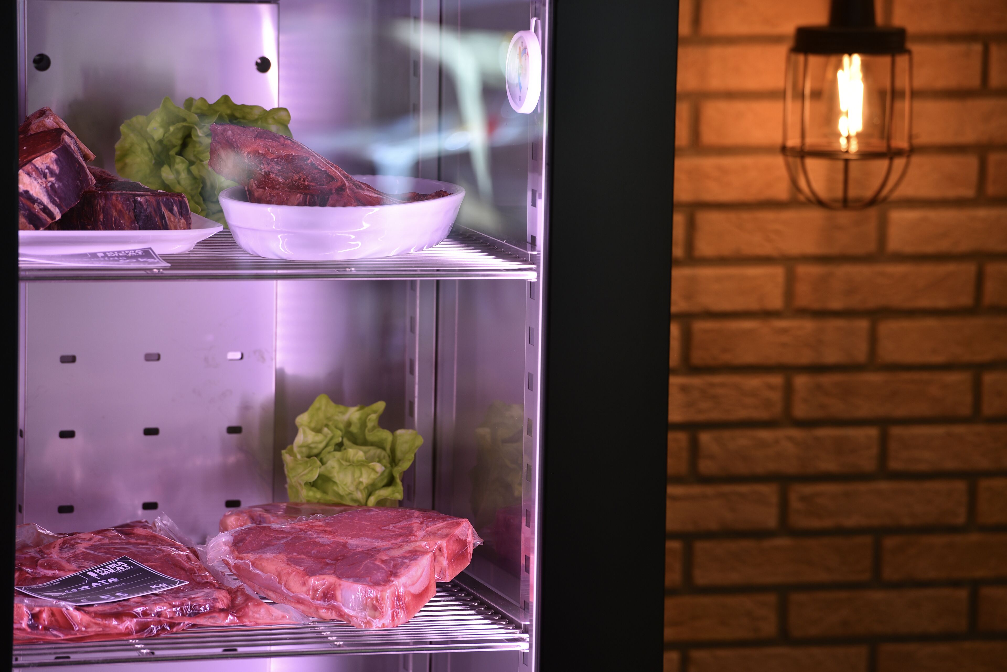 Mostrador refrigerado con carne, pared de ladrillo y lámpara colgante.
