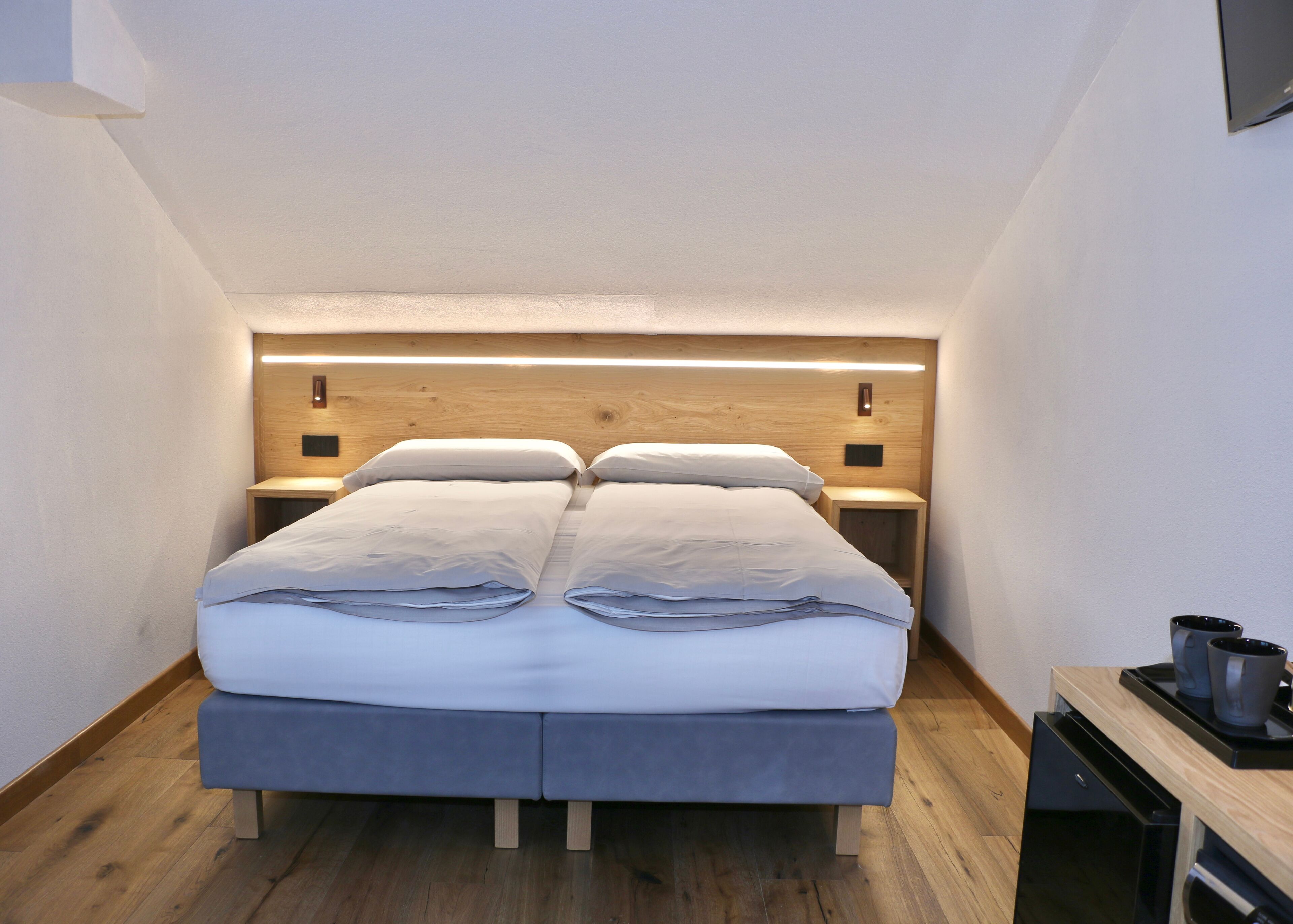 A modern hotel room with a large bed, wooden headboard, bedside tables, and a small desk with a coffee maker.