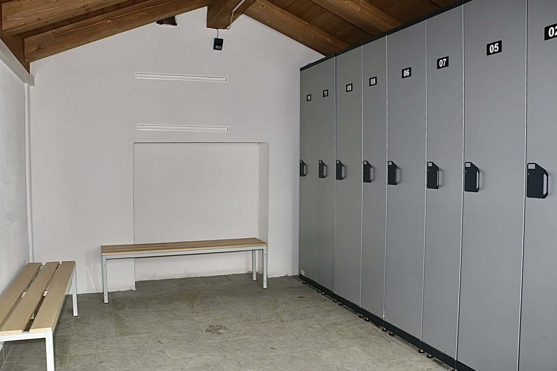 A locker room with grey lockers numbered 05 to 11, a wooden bench, and a white wall with a recessed light fixture.