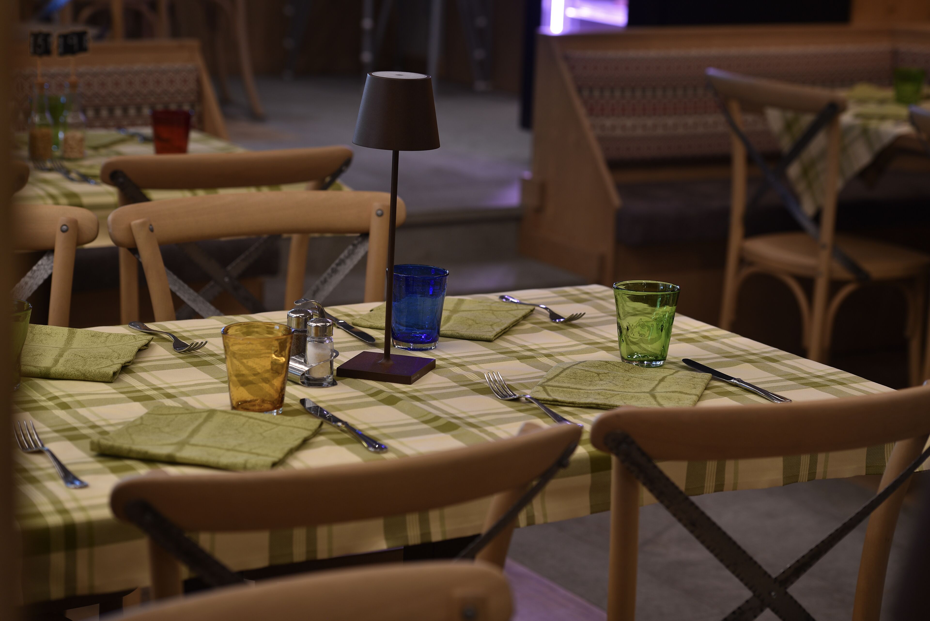 A restaurant setting with checkered tablecloths, wooden chairs, and a lamp on the table.