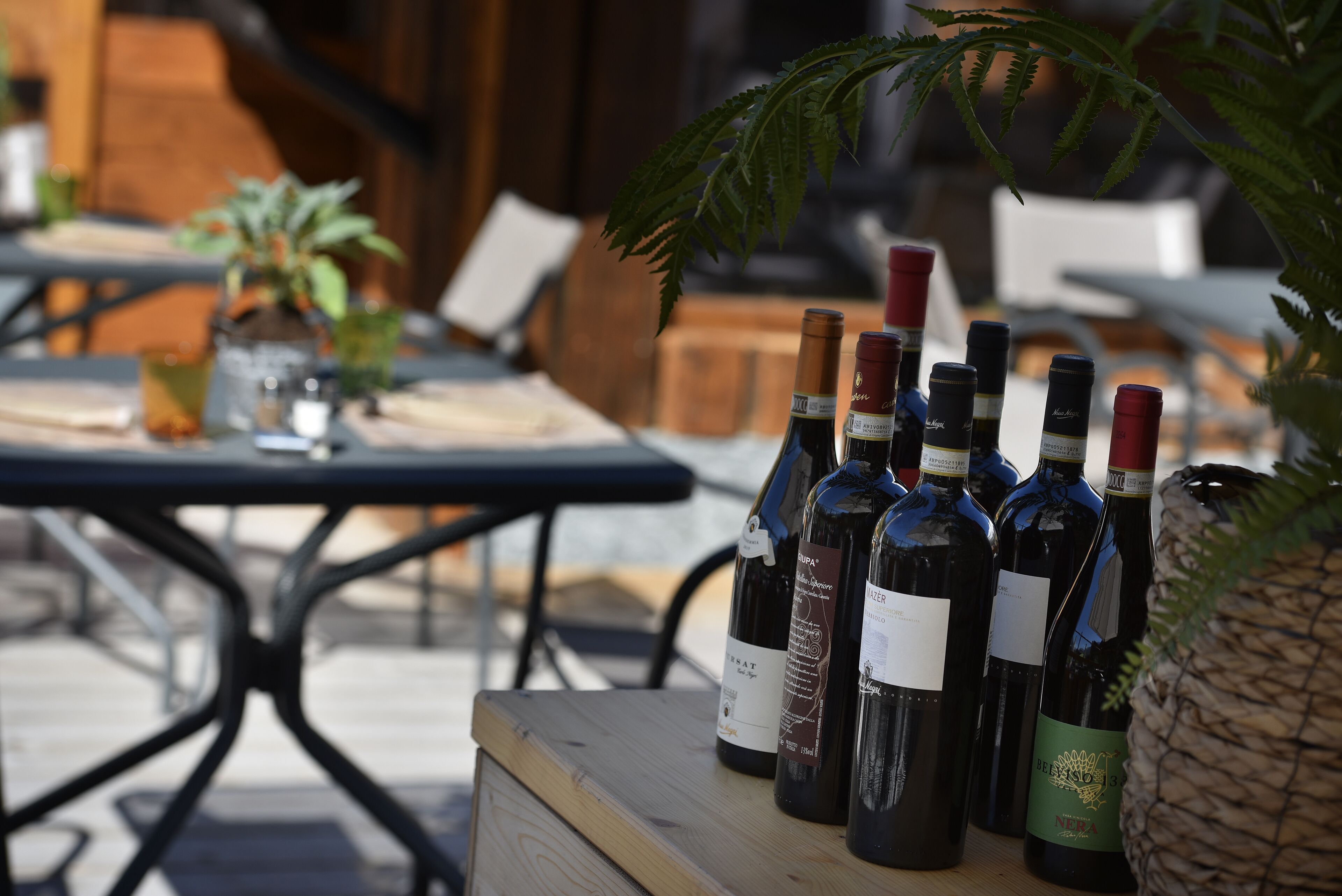 Una mesa de madera con varias botellas de vino y copas, colocada al aire libre con un fondo borroso de mesas y sillas.