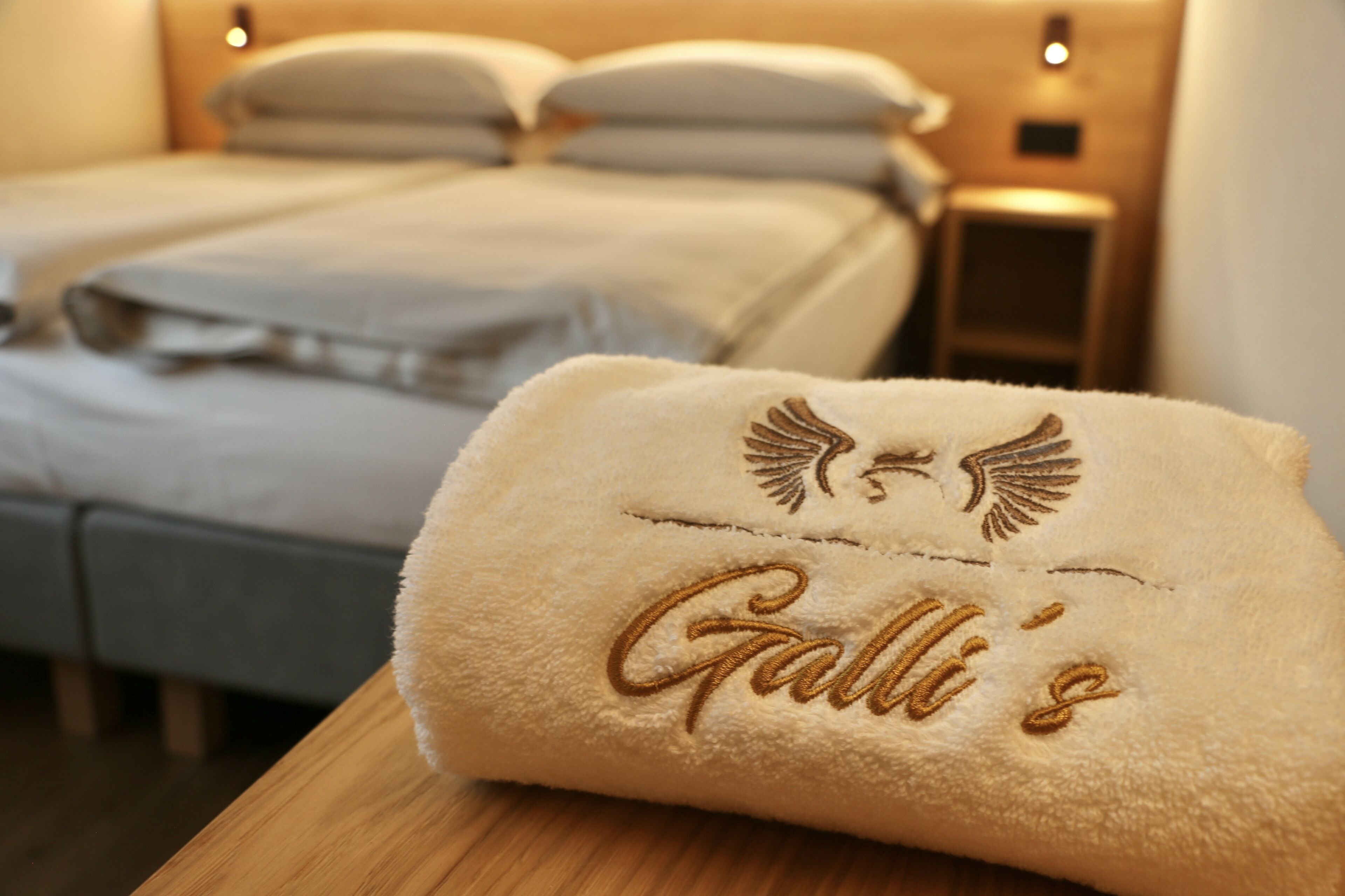 Una cama bien hecha con edredón blanco y almohadas, una mesita de noche de madera con una lámpara, y una toalla con texto bordado y un logotipo alado.