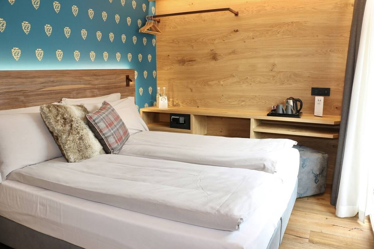 A double bed with white linens, a wooden headboard, a bedside table with a kettle and cups, and a wall with a leaf pattern.