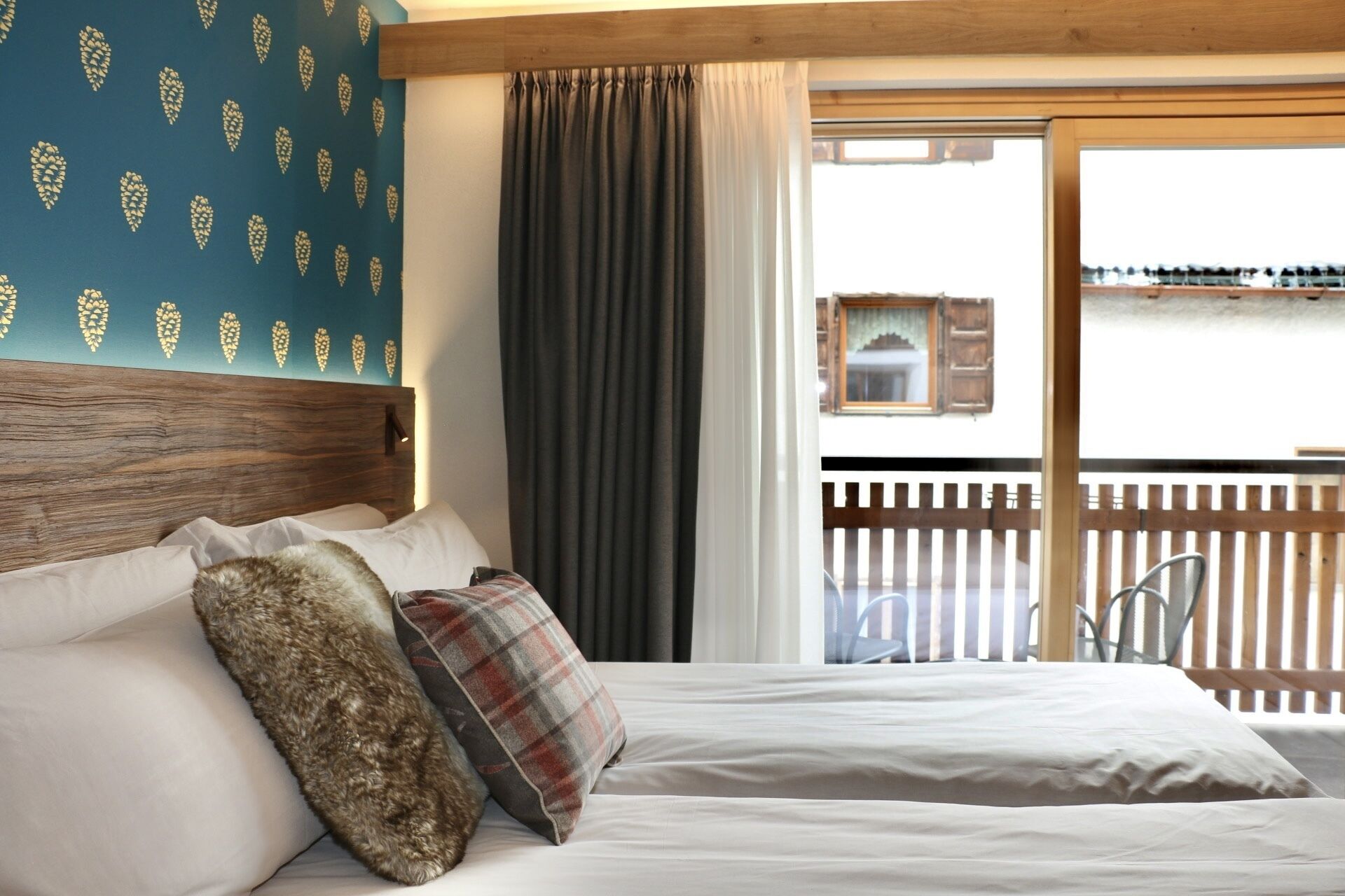 Une chambre à coucher avec une tête de lit en bois, un lit recouvert de draps blancs, un coussin à carreaux et une grande fenêtre donnant sur un balcon et un paysage extérieur.