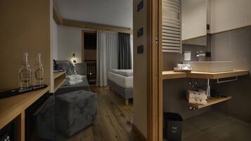 A compact hotel room with a wooden floor, a bed, a small seating area, a kitchenette, and a bathroom with a shower.