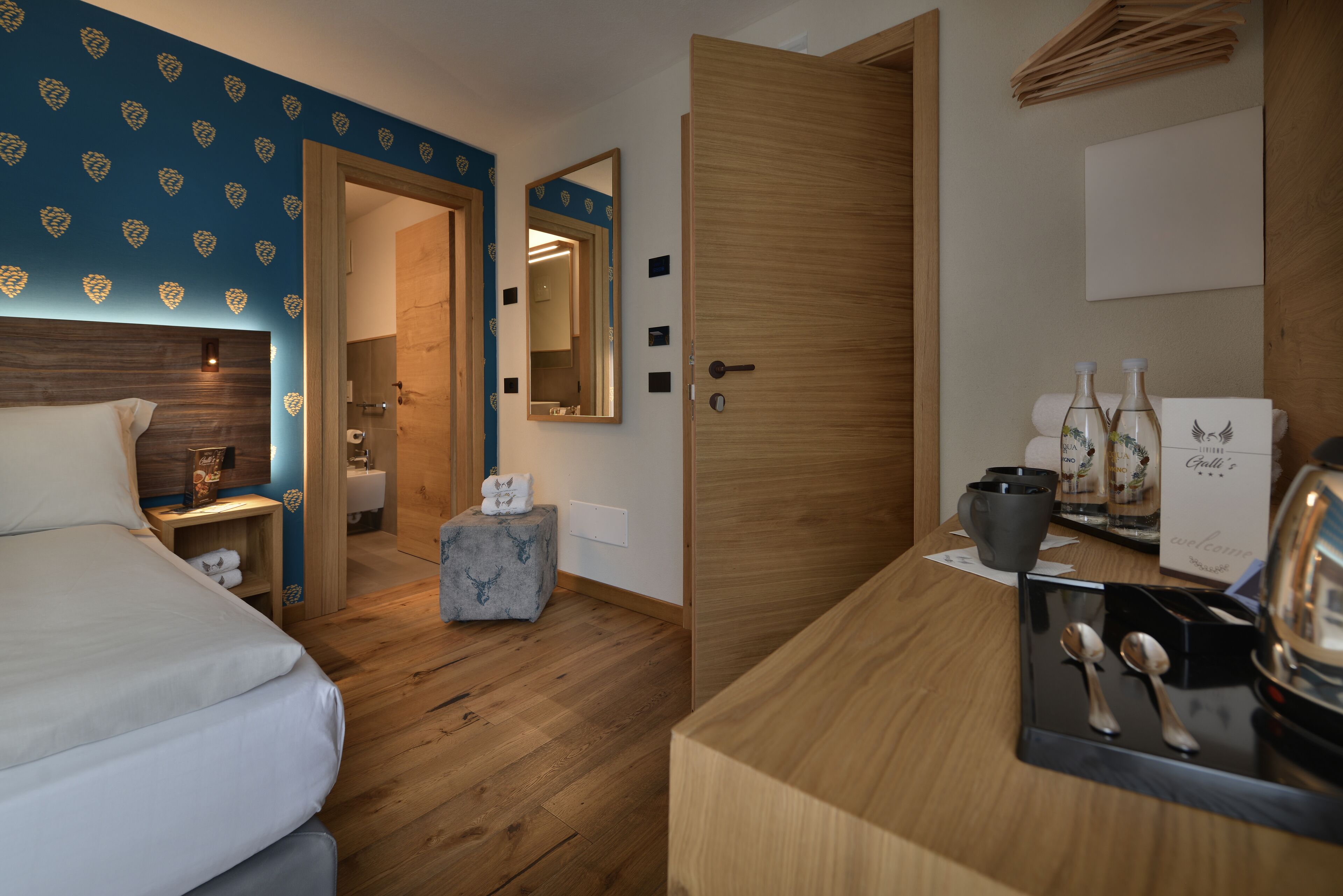 Habitación de hotel con piso de madera, cama, un taburete pequeño, escritorio con espejo y puerta que da a un baño.