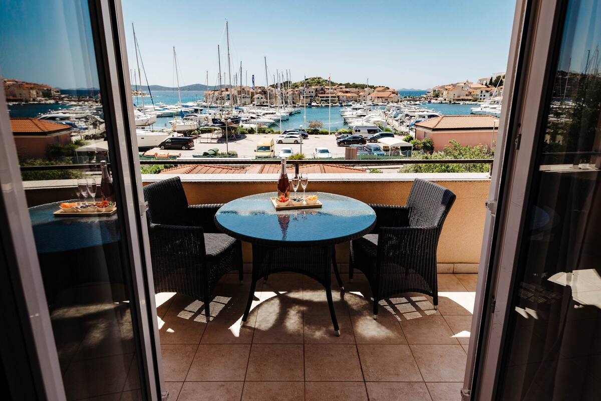 A balcony with a round table and chairs, overlooking a marina with boats and buildings.