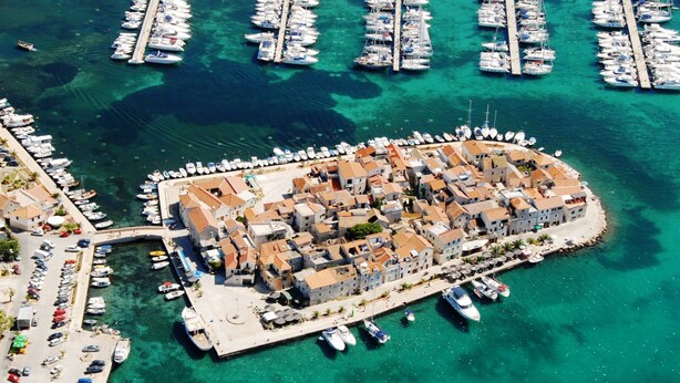 An aerial view of a coastal town with a marina, buildings with red-tiled roofs, and clear turquoise waters. - Villa Diana (Tribunj)