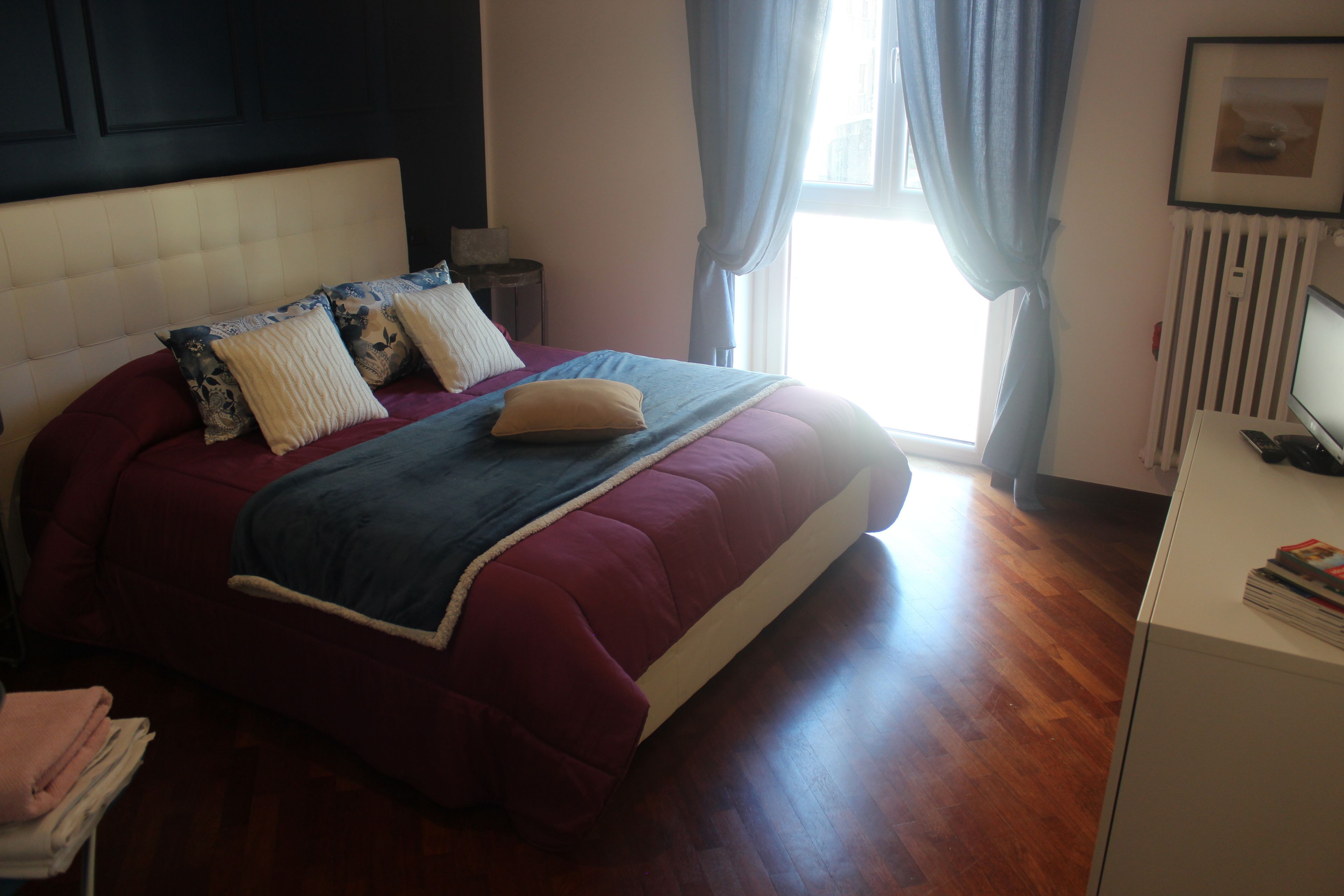 A neatly made bed with a red and blue bedspread, a white headboard, and a bedside table with a lamp.