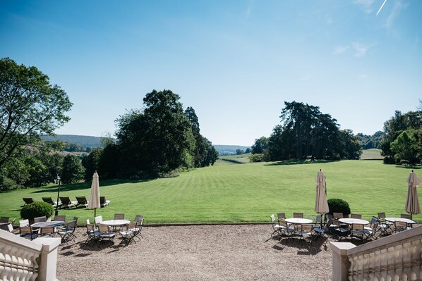 A patio area with tables and chairs, overlooking a well-manicured lawn and trees.