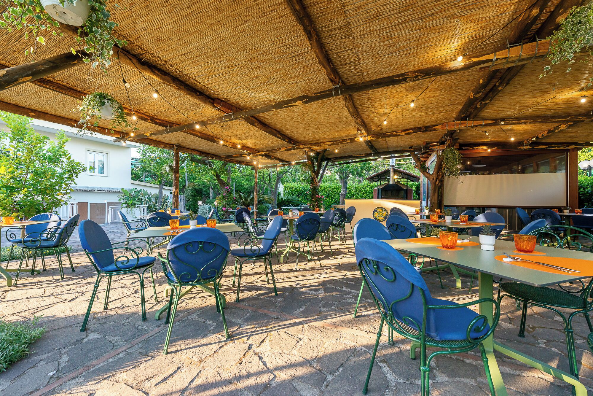 Un espacio de comedor al aire libre con mesas y sillas, techo de paja y plantas colgantes.
