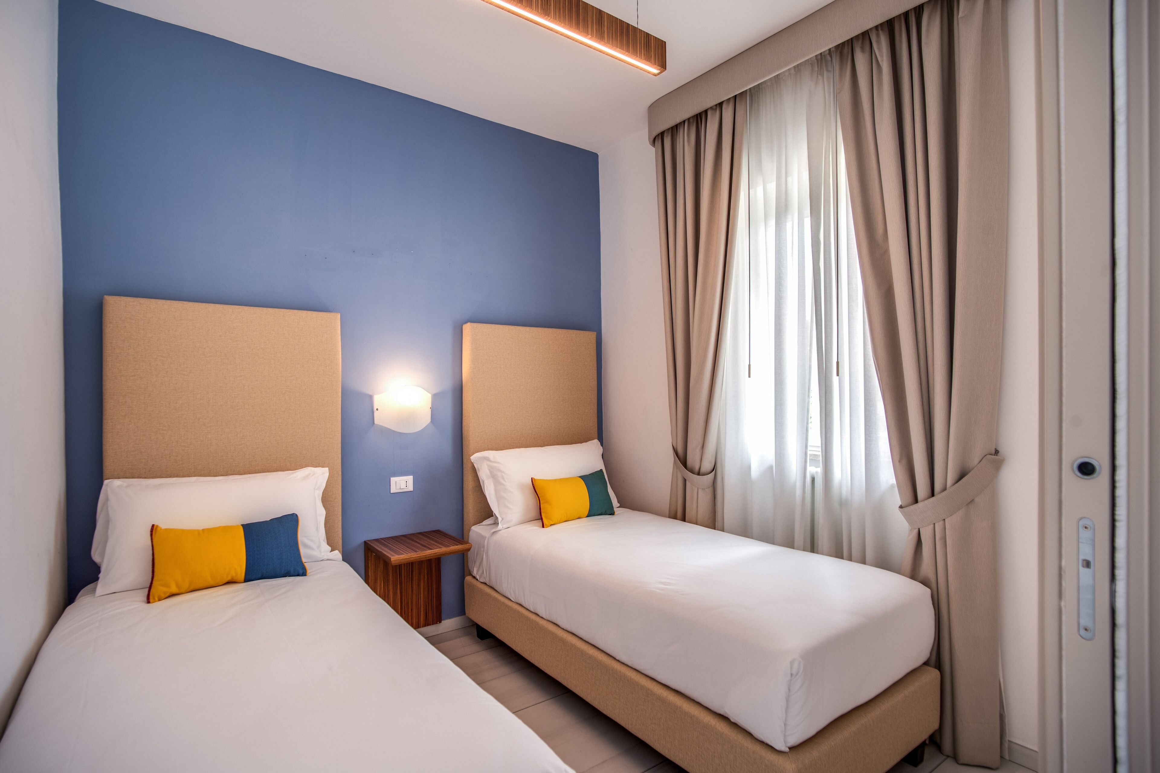 A hotel room with two beds, a nightstand, and a wall-mounted light.