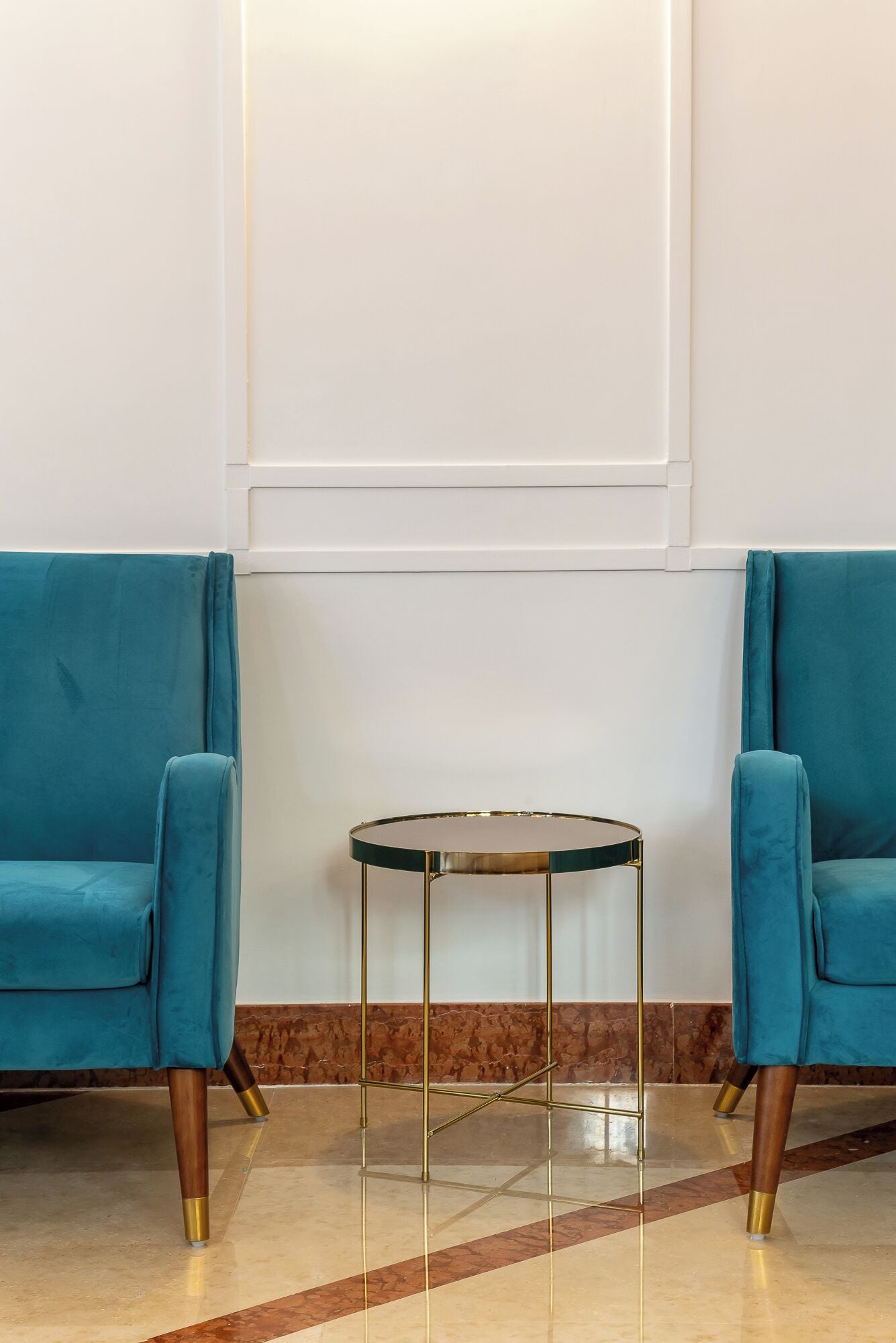 Two teal armchairs, a small round table with a glass top and gold legs, and a marble floor with a brown border.