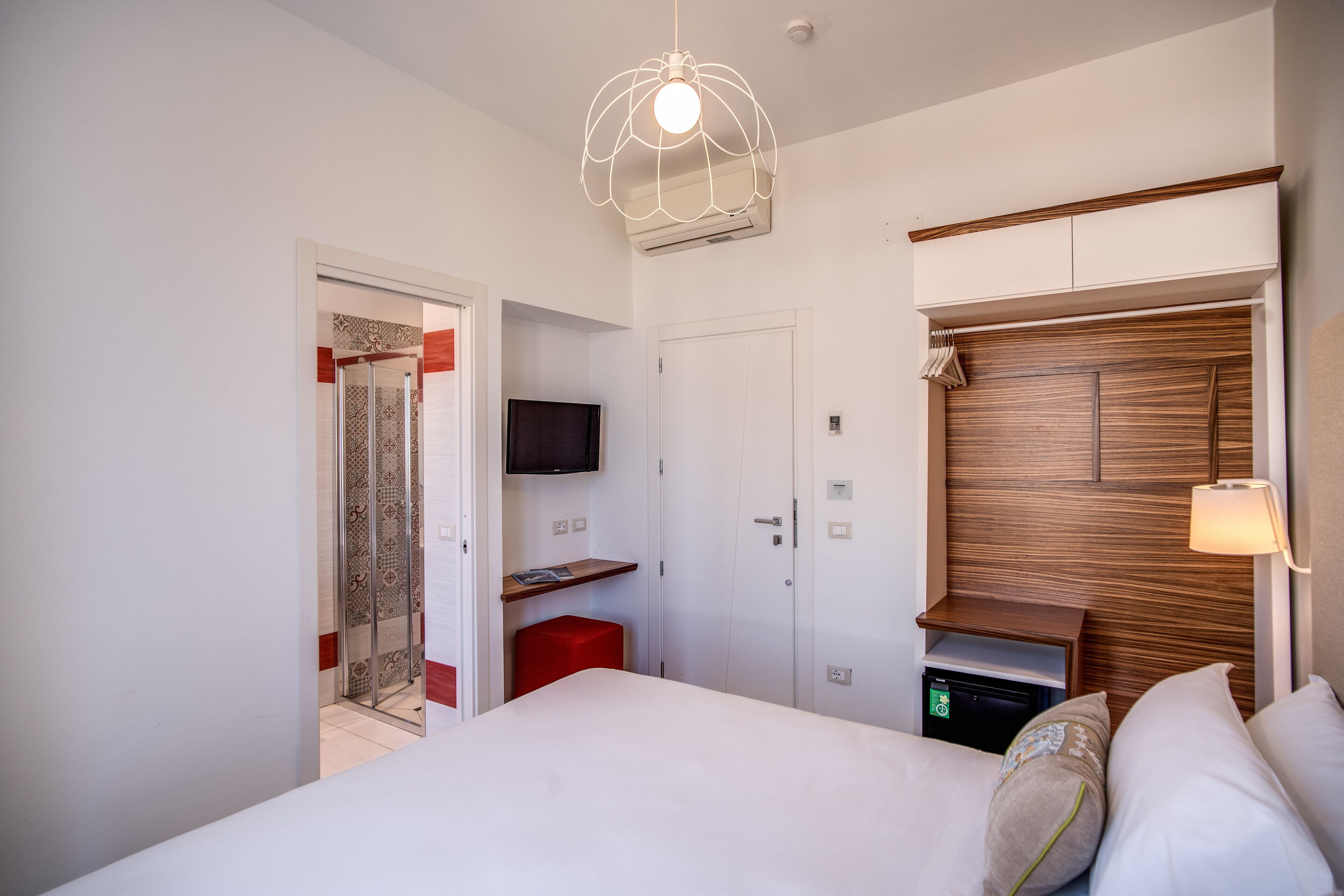 A modern hotel room with a large bed, a TV mounted on the wall, a wooden headboard, and a red accent wall.