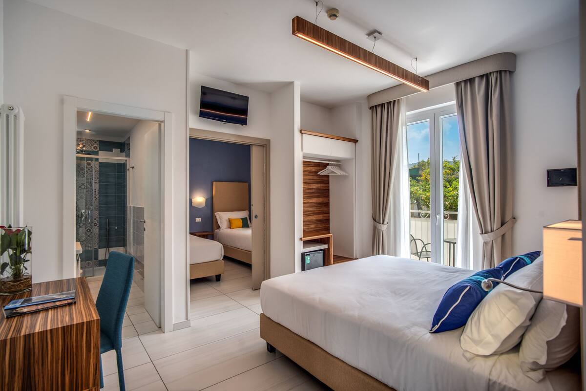 A modern hotel room with a large bed, a desk, and a seating area. There is a balcony with a view of the outdoors.