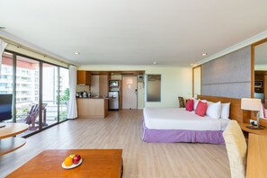 A hotel room with a bed, a desk, a dining table with fruit, a kitchen area, and large windows. - Bella Villa Metro (Pattaya)