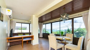 A spacious room with a dining area, a sofa, and a large window offering a view of the outdoors. - Bella Villa Metro (Pattaya)