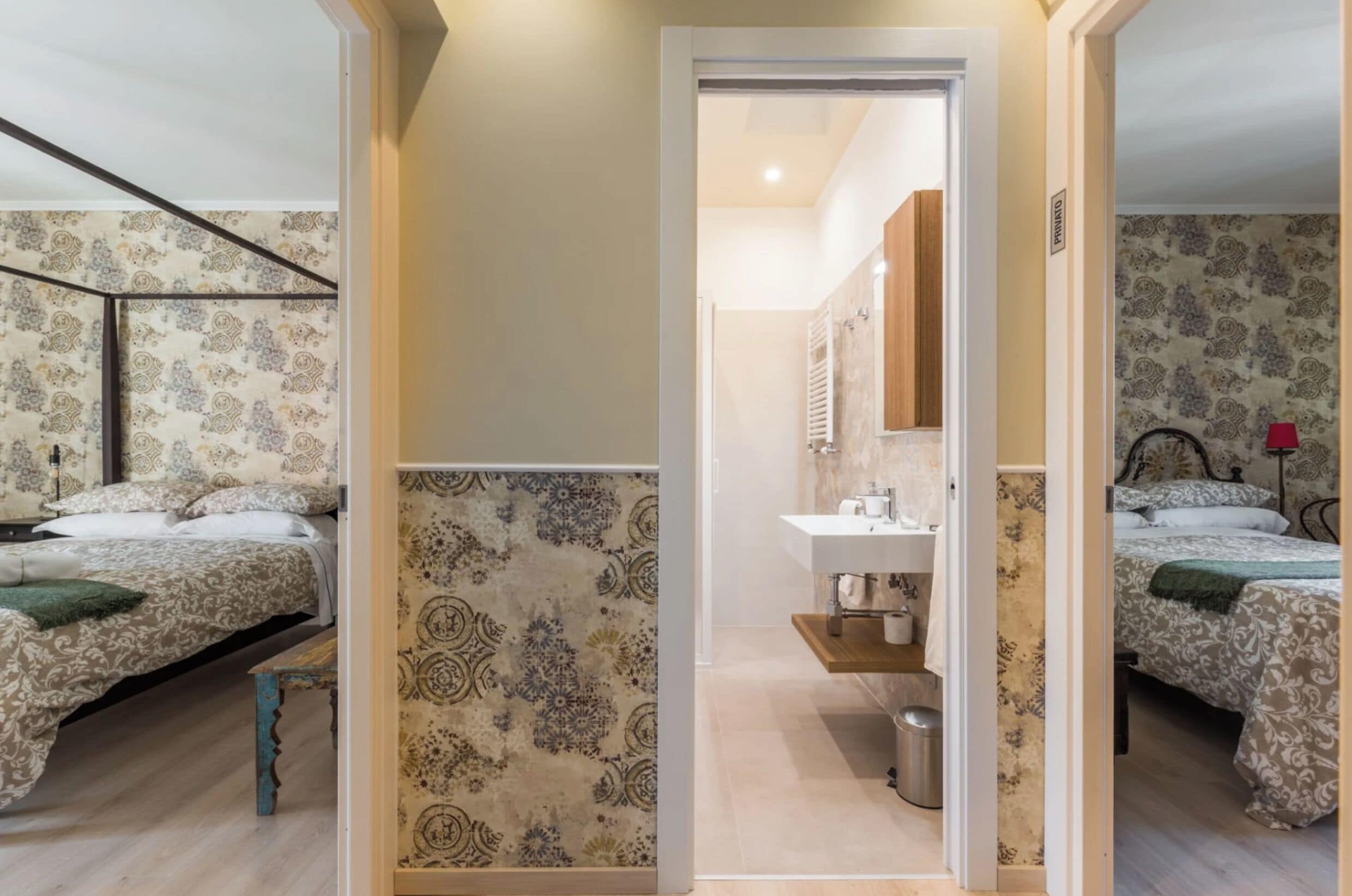 A room with two beds, a bathroom with a sink, and patterned wallpaper.