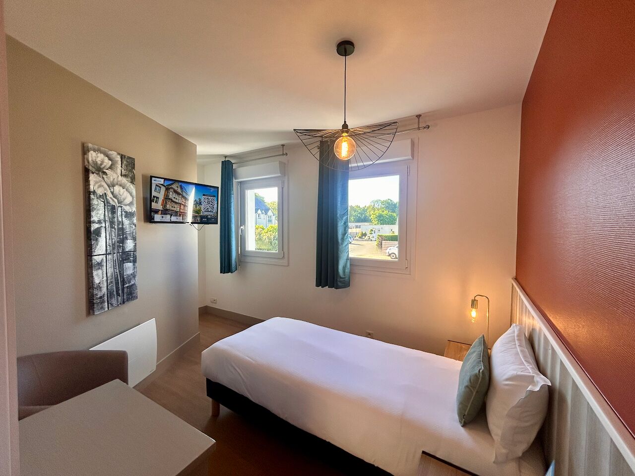Ein Schlafzimmer mit einem Bett, einem Schreibtisch, einem Stuhl, zwei Fenstern mit Vorhängen und einer Wandleuchte.