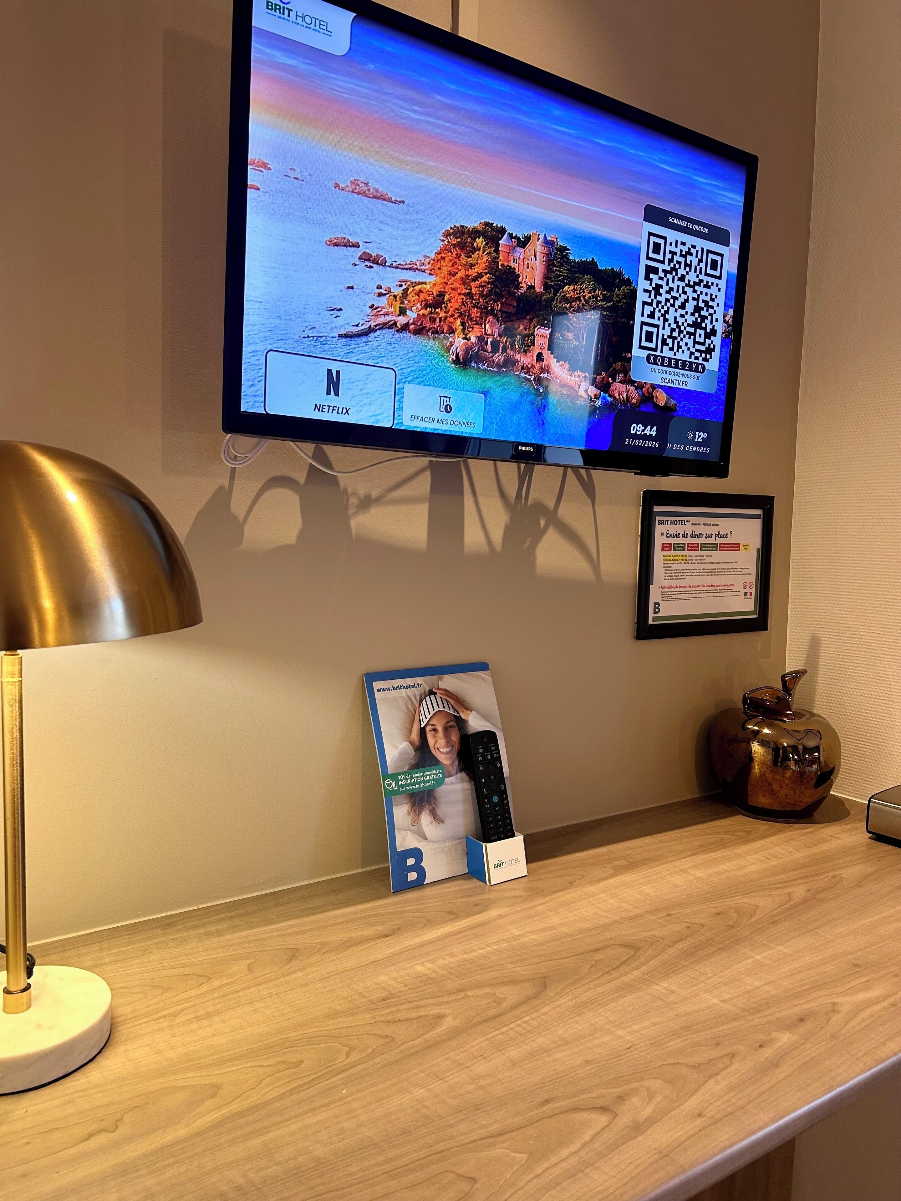 A flat-screen TV mounted on a wall displaying a scenic coastal image, a framed certificate, a remote control box, and a lamp on a wooden desk.