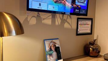 A flat-screen TV mounted on a wall displaying a scenic coastal image, a framed certificate, a remote control box, and a lamp on a wooden desk.
