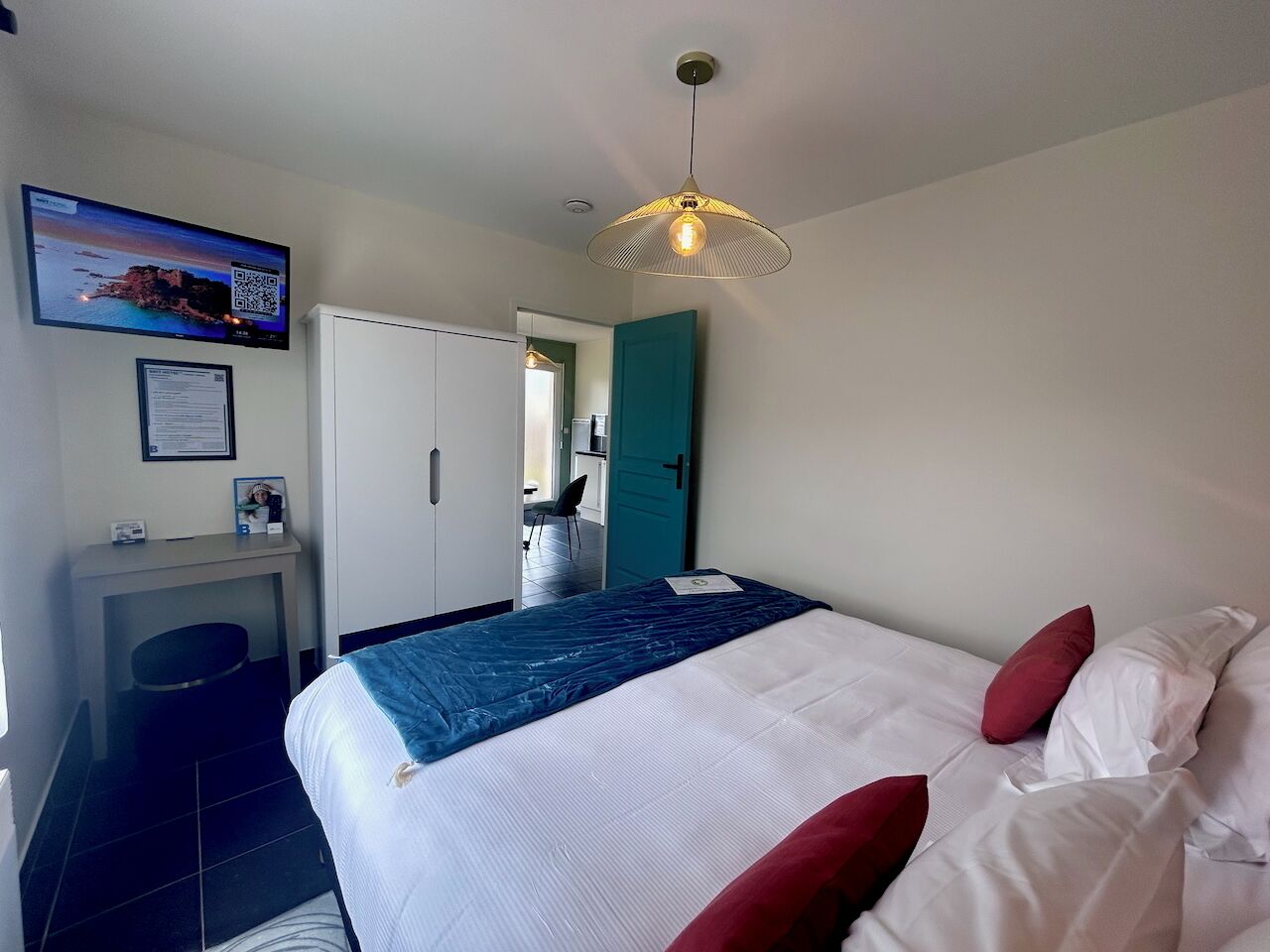 Een hotelkamer met een bed, een bureau met stoel, een aan de muur bevestigde televisie en een deur naar een andere kamer.