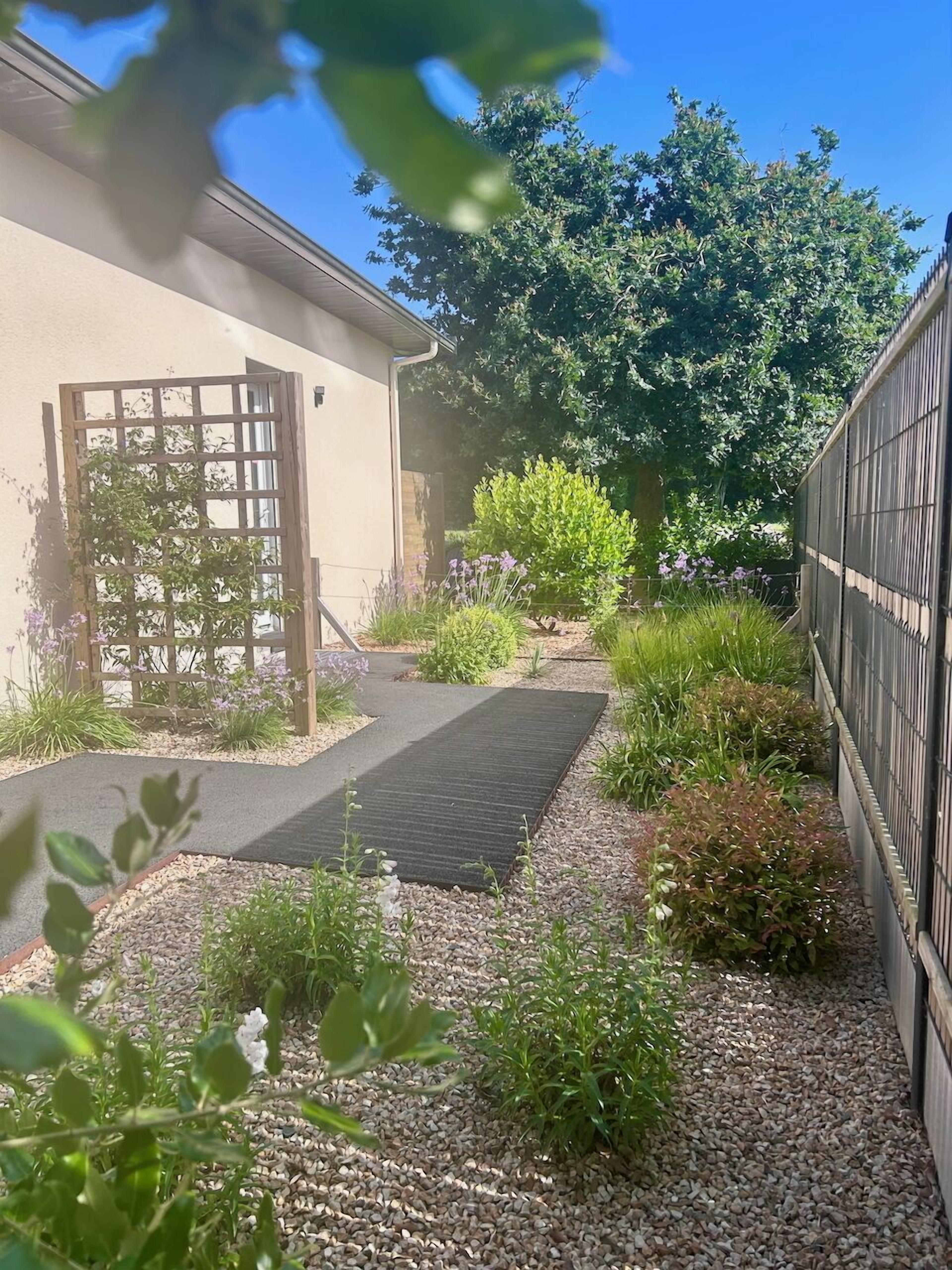 Un chemin de jardin menant à une maison dotée d’un portail en bois, entourée de gravier et de différentes plantes.
