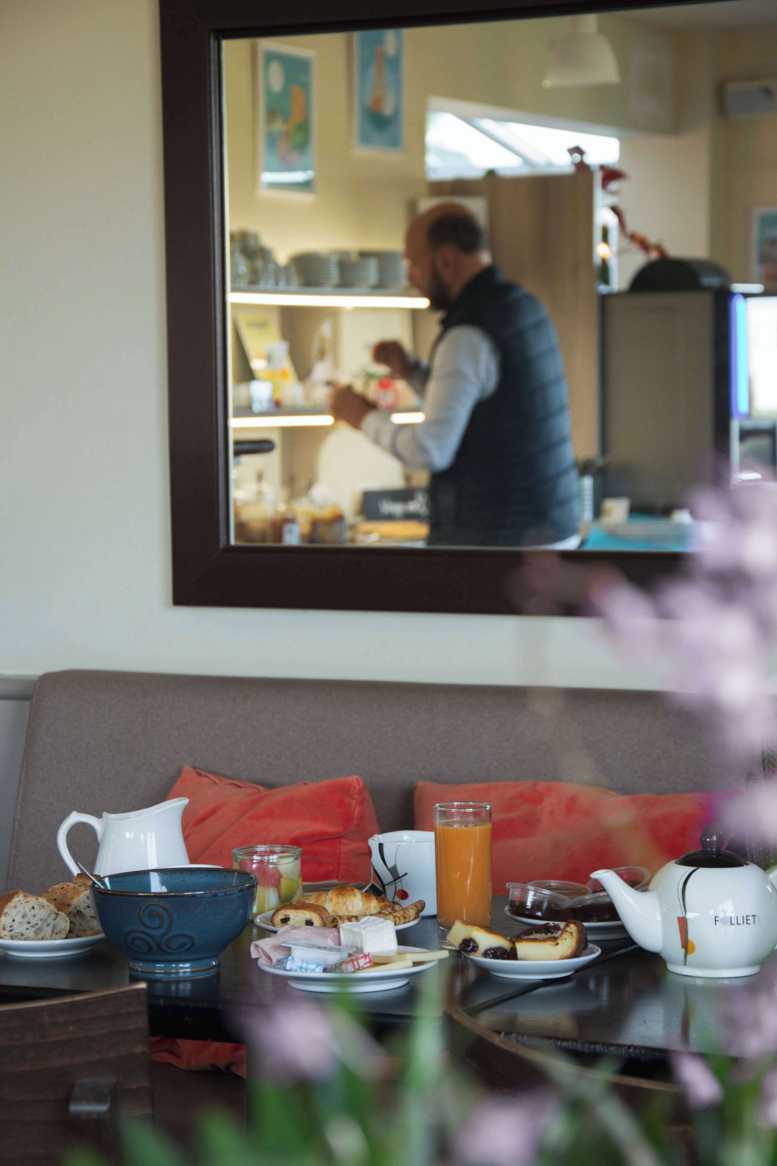A breakfast table with a pitcher, bowl, and teapot, and a mirror reflecting a person in the background.