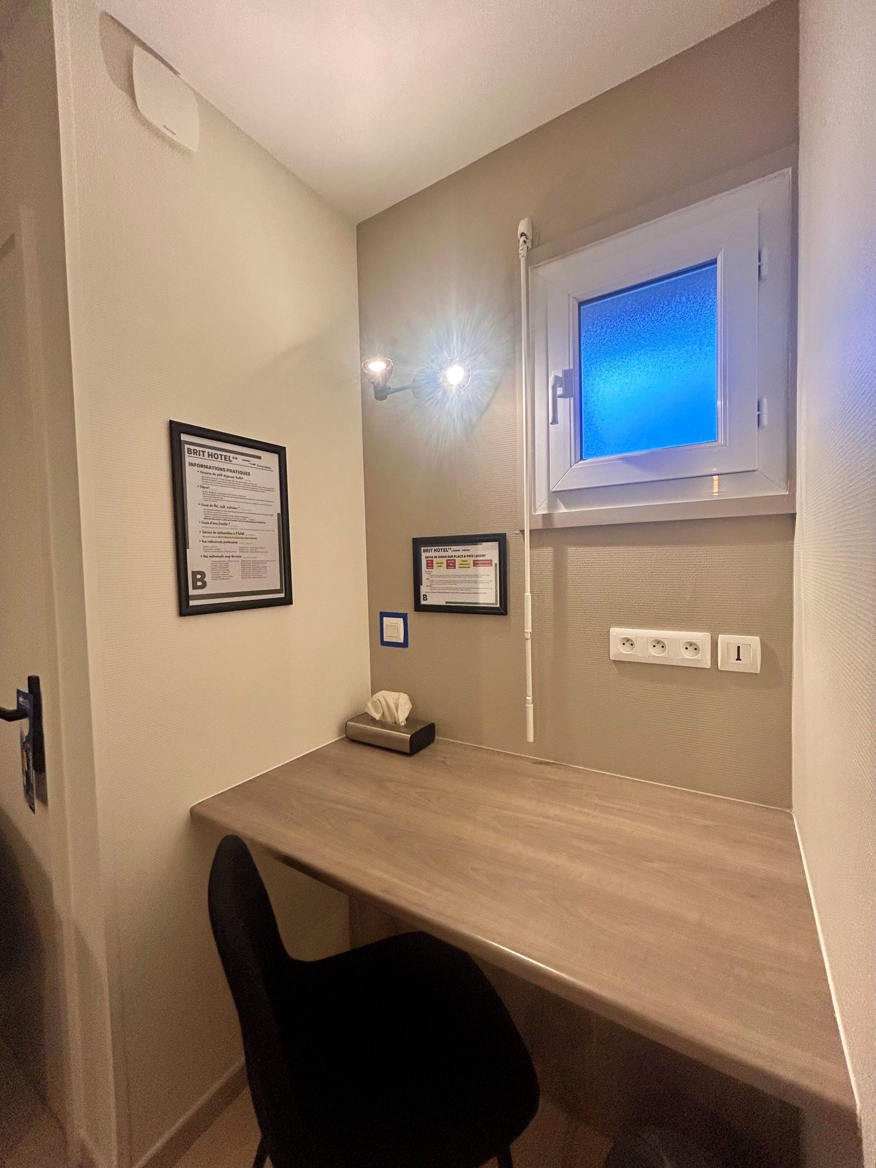 A hotel room with a desk, a black chair, a window with a view, and wall-mounted lights.