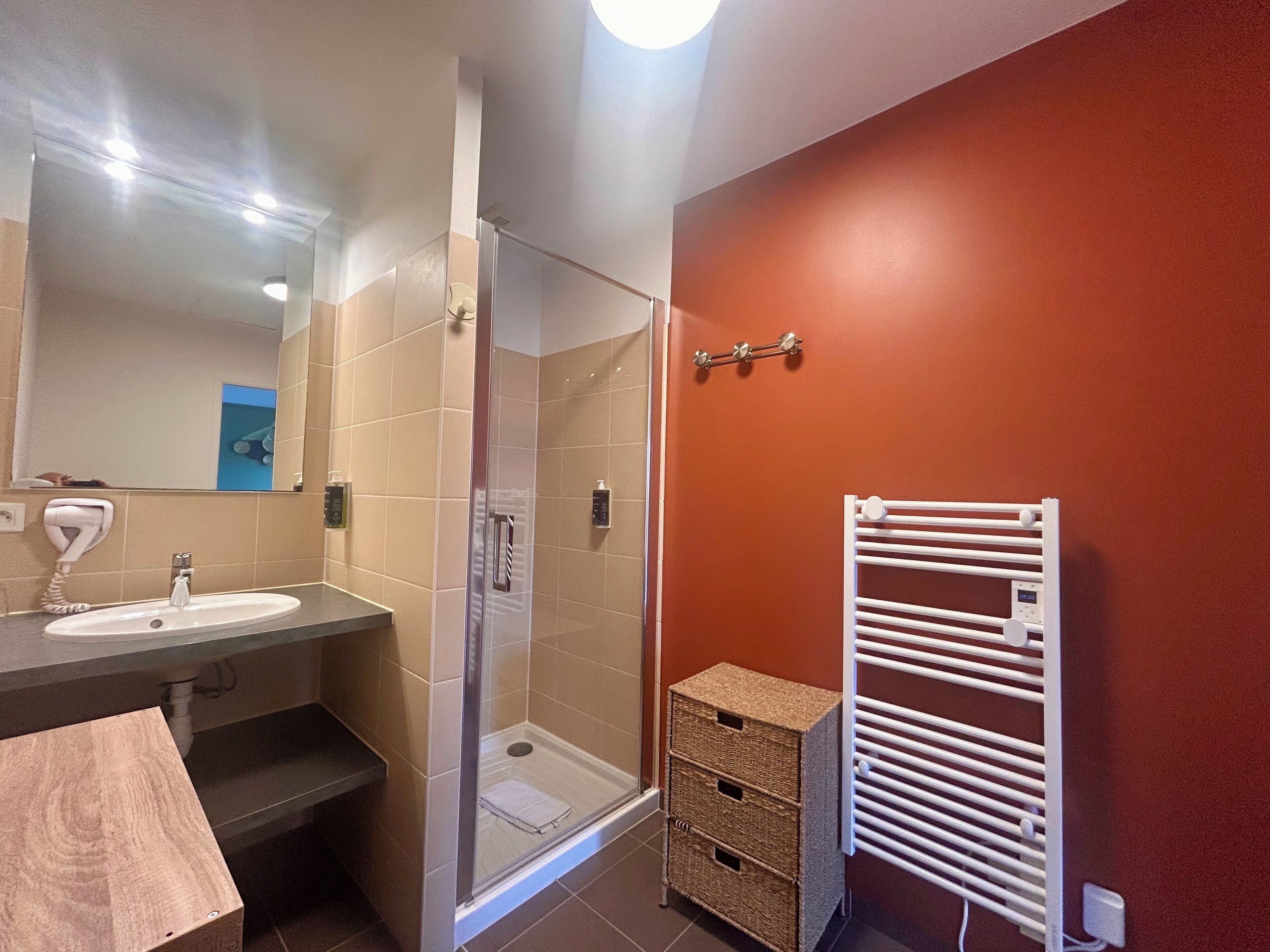 A bathroom with a glass shower enclosure, a sink with a hairdryer, a towel rack, and a small wicker basket.
