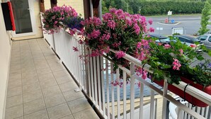 Ein Balkon mit weißer Brüstung, Blumentöpfen mit rosa Blüten und Blick auf einen Parkplatz und Bäume im Hintergrund.