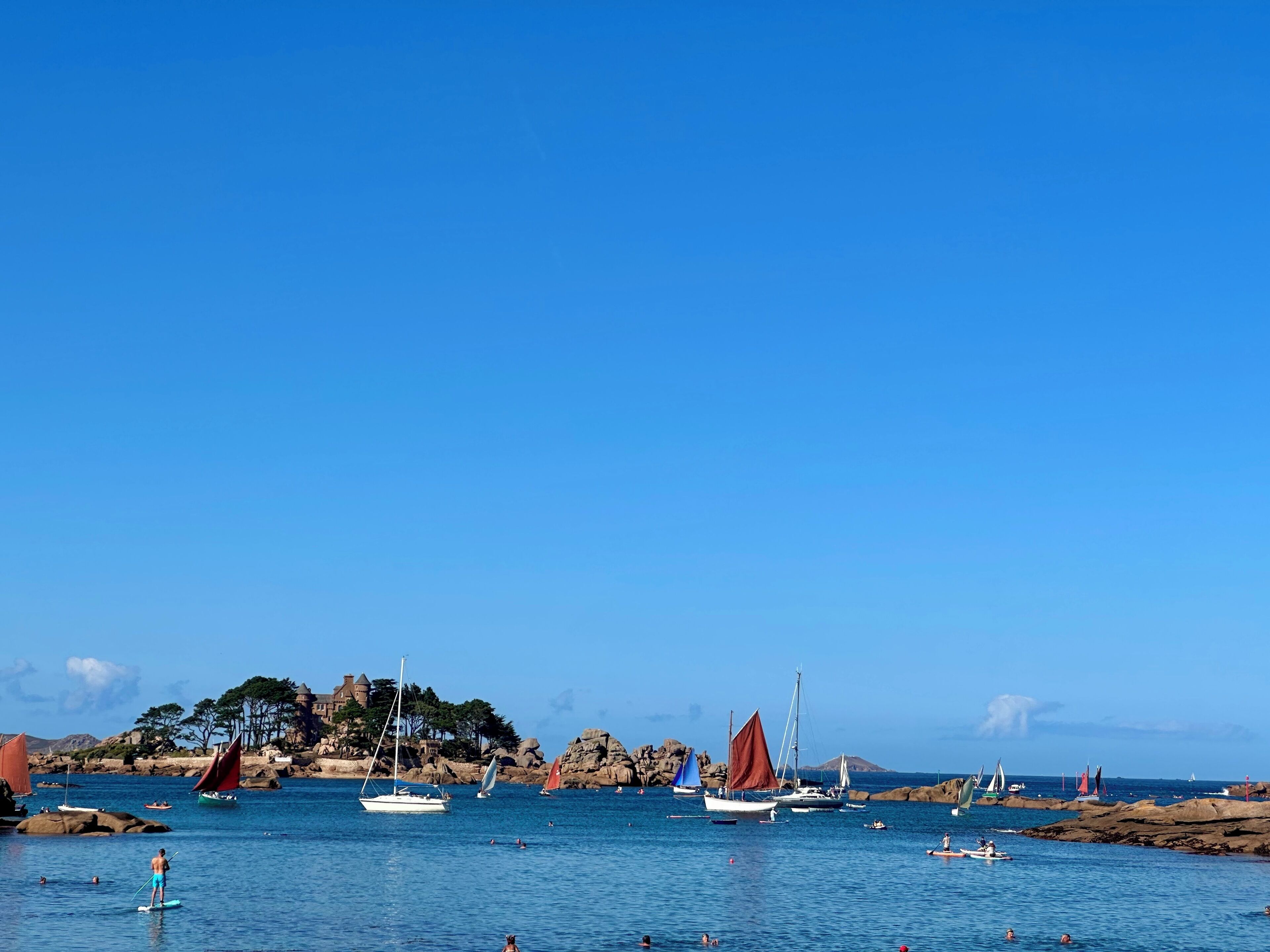 A coastal scene with boats, people swimming, and a rocky shoreline with buildings.