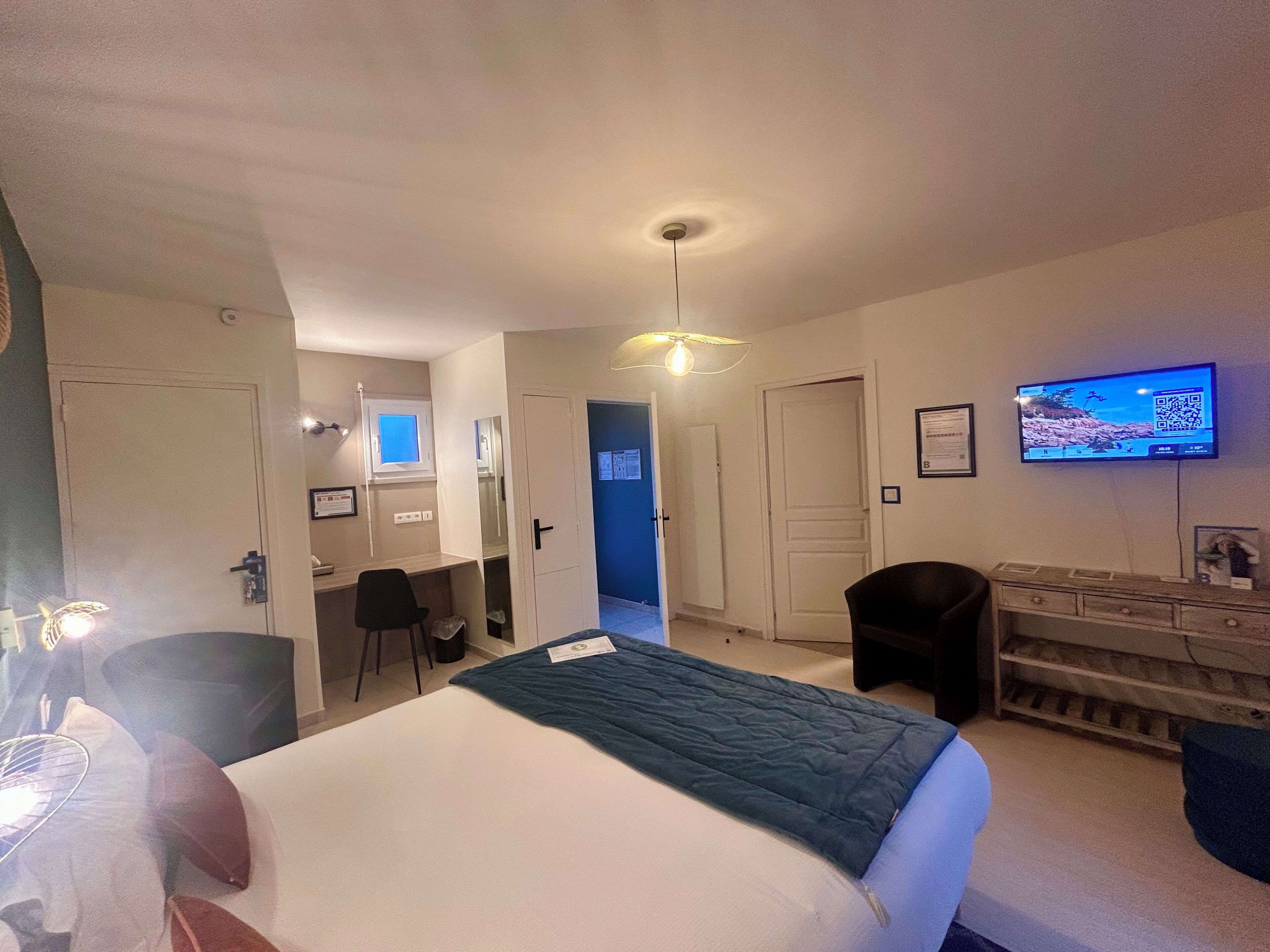 Quarto de hotel com cama, escrivaninha, cadeira, televisão e guarda-roupa.