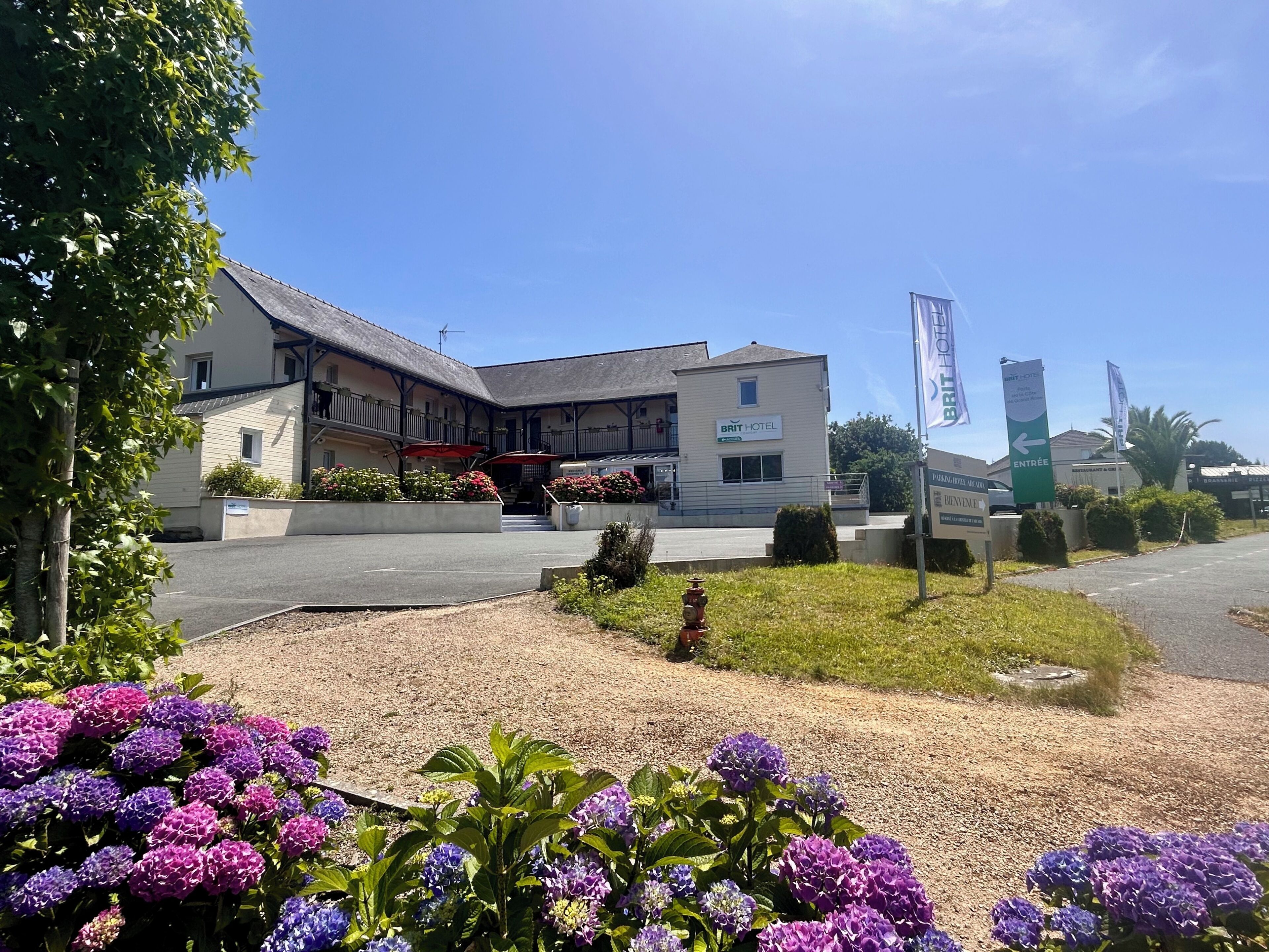 Un hôtel doté d’un espace extérieur couvert avec des sièges, d’une enseigne indiquant qu’il s’agit d’un hôtel de la chaîne Brit Hotel, et d’un parking.