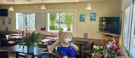 A restaurant interior with a large teddy bear, dining tables, chairs, potted plants, a menu board, and a TV.