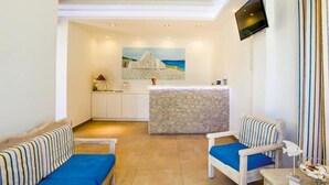 A room with a stone wall, a reception desk, a TV mounted on the wall, and striped furniture. - Anemos Studios & Apartments (Mykonos)
