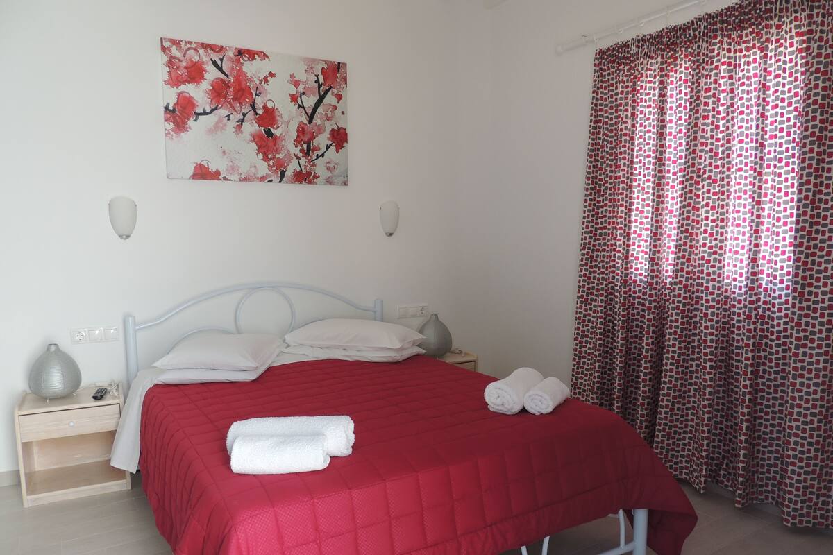 Una cama bien hecha con colcha roja, almohadas blancas y dos toallas enrolladas a un lado.