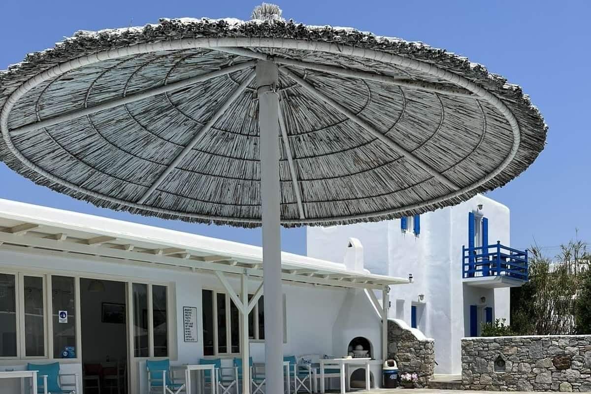 Un área de piscina con una sombrilla de paja, sillas de descanso y un edificio blanco con detalles azules.