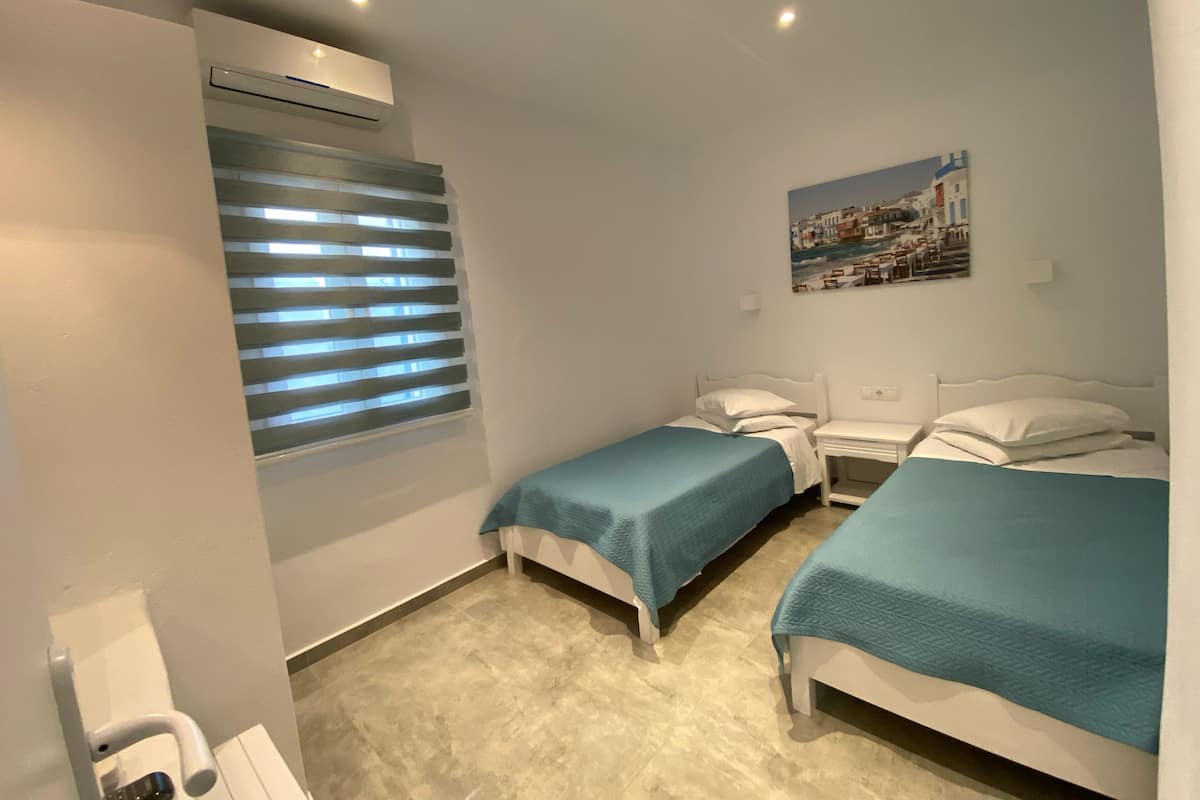 Una habitación de hotel con dos camas individuales, una ventana con persianas, un equipo de aire acondicionado, una mesita y un cuadro colgado en la pared.