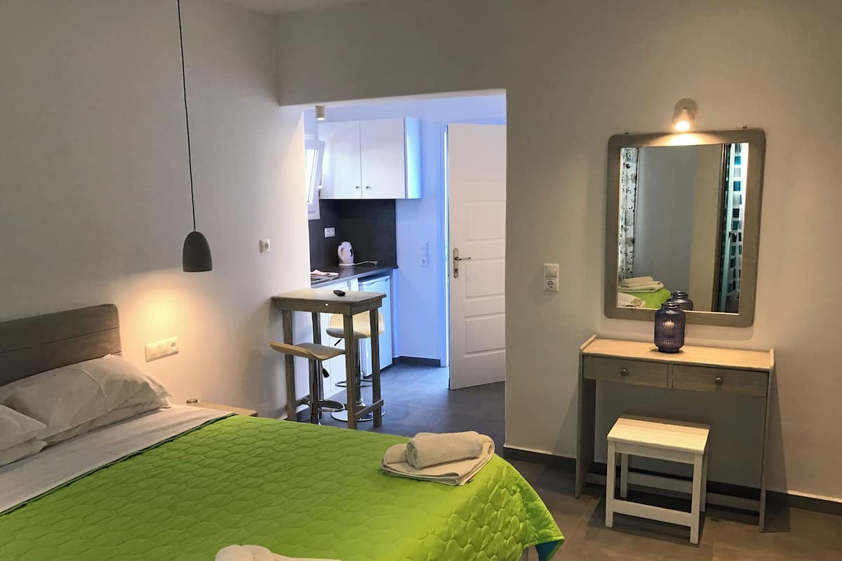 Habitación de hotel con una cama verde acolchada, una mesita de noche con espejo, un taburete pequeño y un banco. Se ve una kitchenette al fondo.