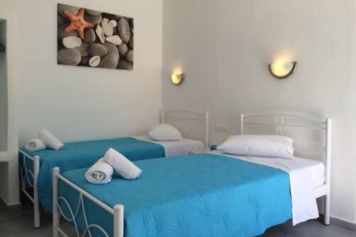 Habitación de hotel con dos camas individuales, cada una con cubrecama azul y almohadas blancas. En la pared, hay una obra de arte con una estrella de mar y piedras.