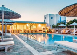 A pool area with lounge chairs and umbrellas, a building with a stone facade, and a clear sky. - Anemos Studios & Apartments (Mykonos)