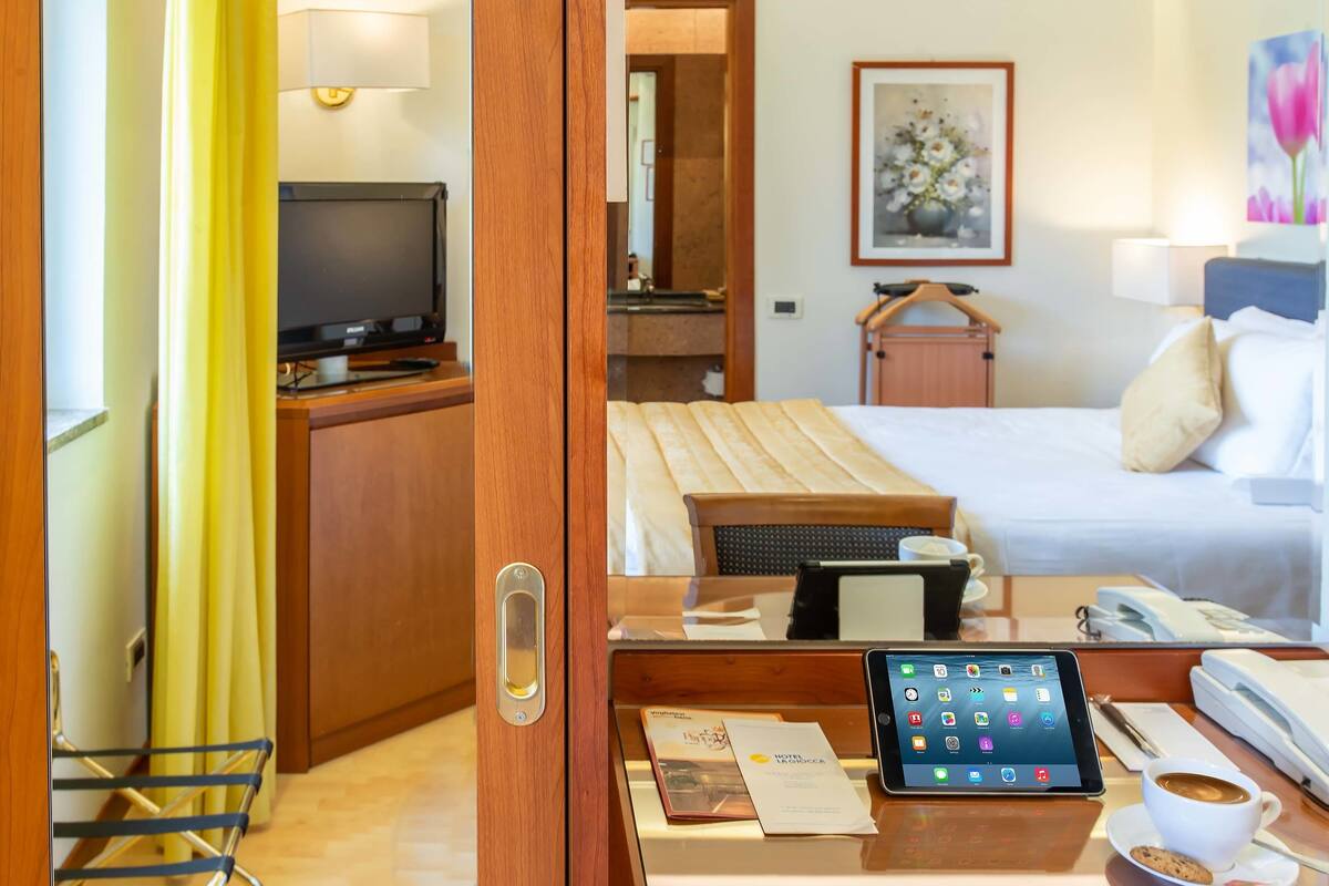 A hotel room with a bed, a desk with a tablet, a coffee cup, a TV, and a bathroom visible through an open door.